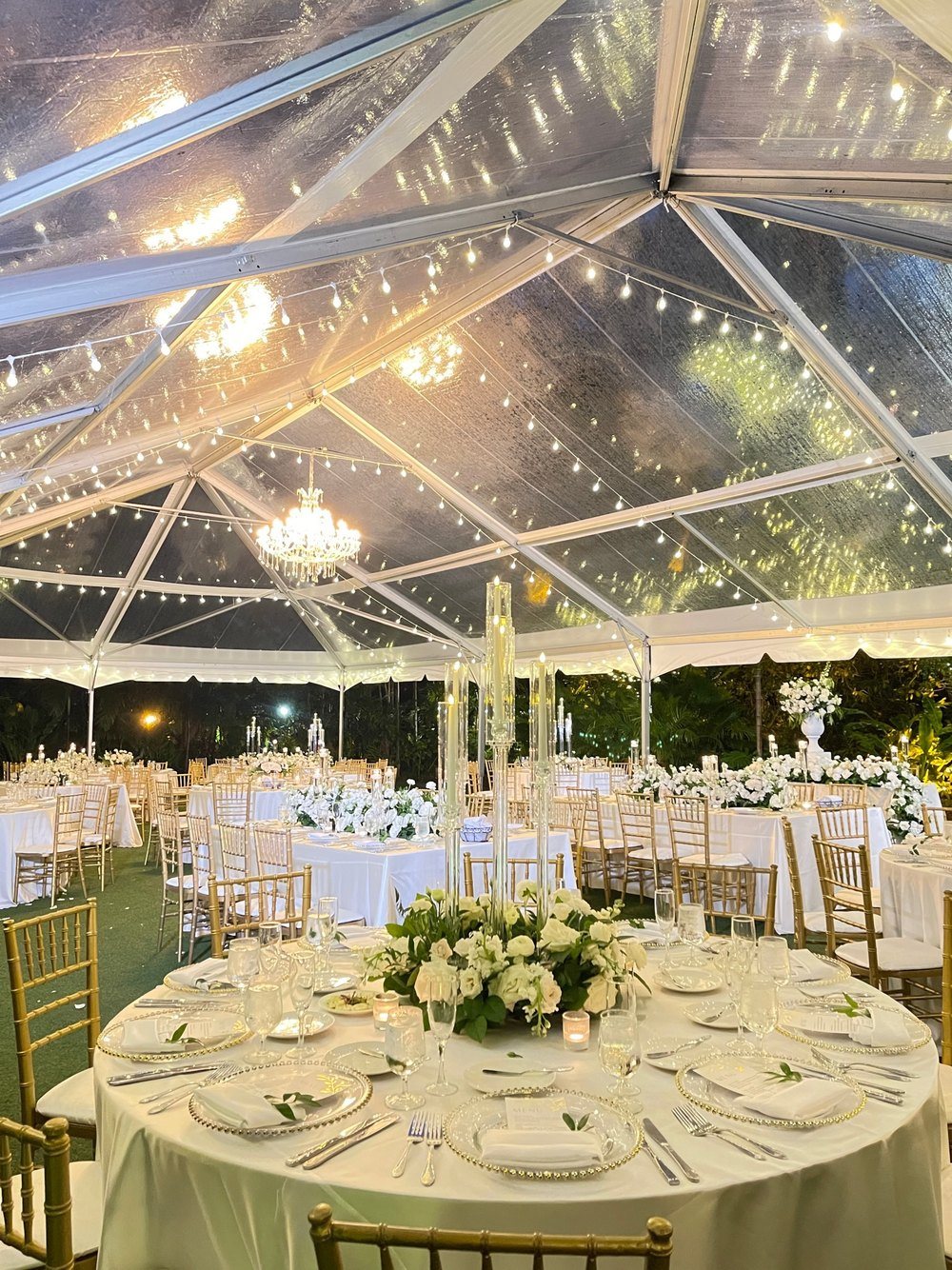 A clear-top tent dinner with a crystal chandelier, string lights, white floral centerpieces and gold chairs at night