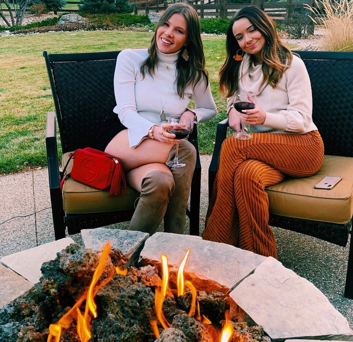 Two guests in cozy sweaters sharing red wine beside an outdoor fire pit at dusk