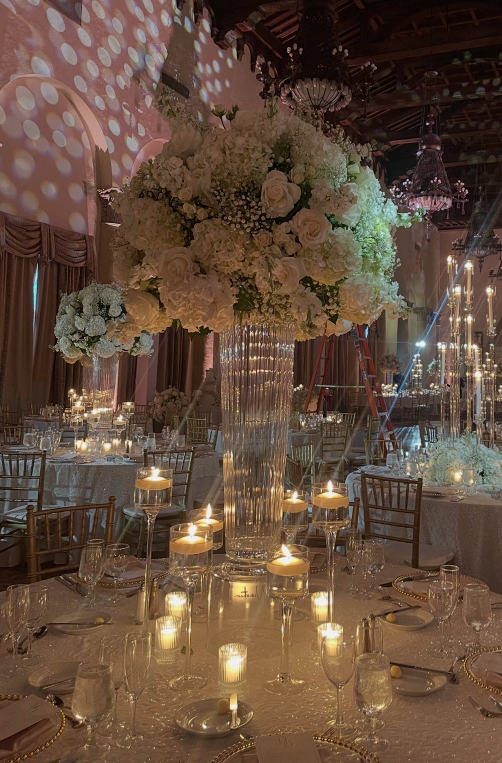 A towering white hydrangea and rose centerpiece in a tall crystal vase over a candlelit reception table